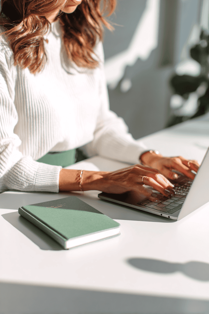 Mother seated at white desk, typing on laptop with a sage green journal beside her