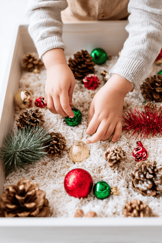 Toddler’s hands scooping and arranging items in a holiday sensory bin, rice base with jingle bells, ribbons, and pinecones visible