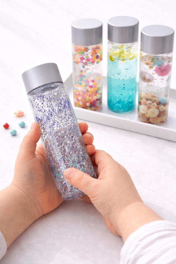 Preschooler holding and tilting a glitter sensory bottle, with other clear sensory bottles filled with beads, oil and water, and small objects arranged on a white tray