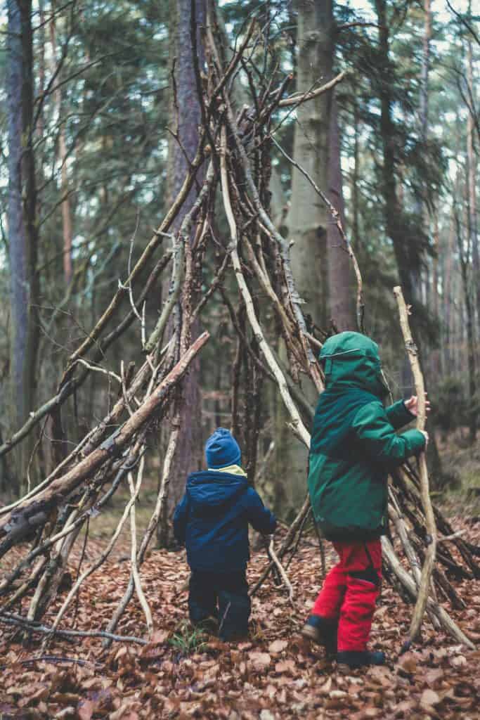two young children exploring a handmade stick fort in a dense, autumn forest