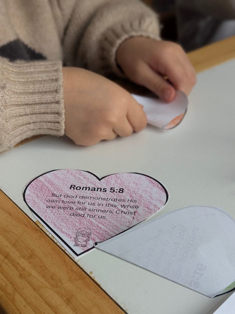 Child’s hands folding a heart-shaped Romans 5:8 Bible verse card for a Jesus Loves Me Christian craft