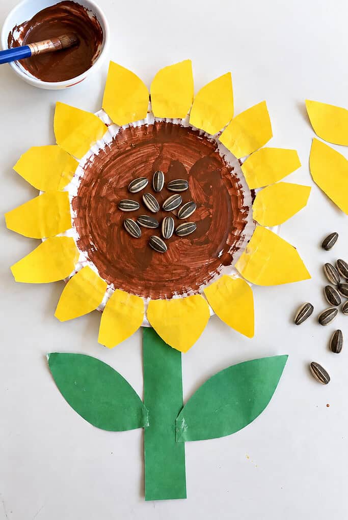 A paper plate sunflower with a brown painted center pressed with real sunflower seeds and a ring of yellow paper petals, with a green paper stem and leaves, surrounded by a glue stick, a dish of brown paint, and loose seeds on a white table.