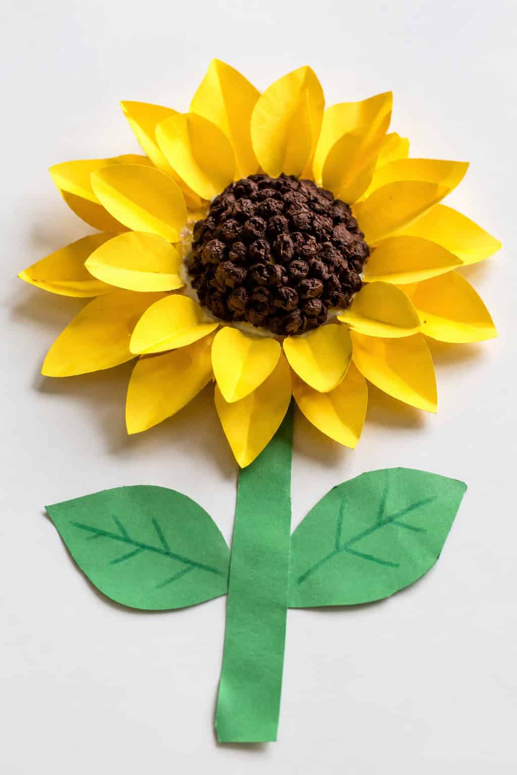 A 3D sunflower craft with folded and curled yellow paper petals standing up in two layered rings around a raised brown tissue center, with a green paper stem and leaves, photographed at an angle on a white table to show its depth.
