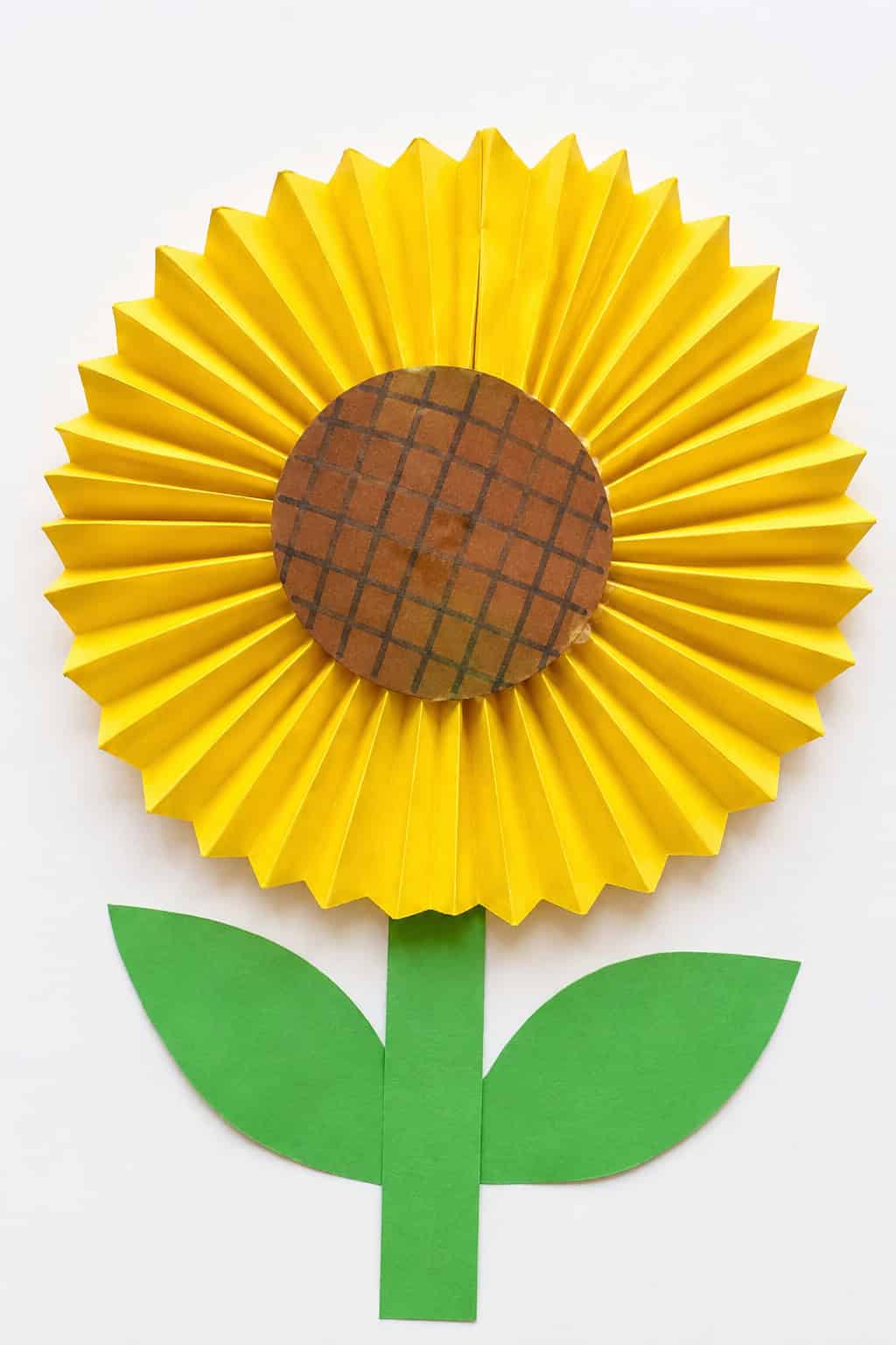 An accordion-fold origami sunflower with yellow pleated fan-folded petals forming a full circle around a brown center drawn with a crosshatch seed pattern, with a green paper stem and leaves on a white table.