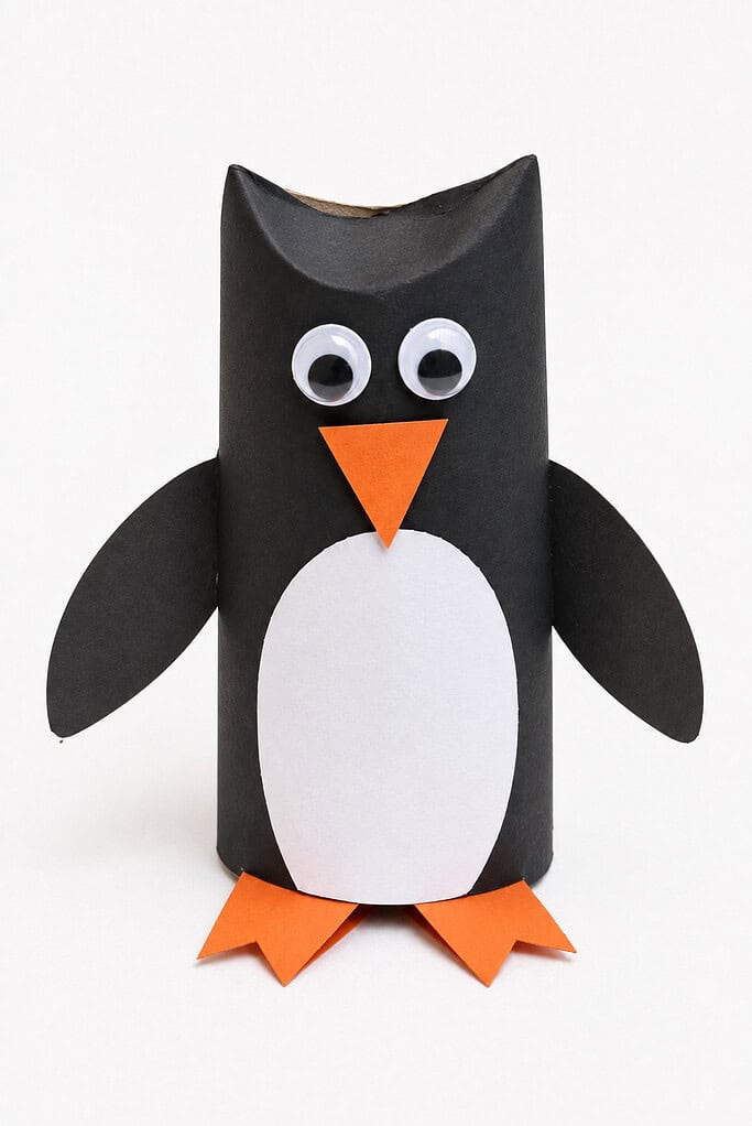 a toilet paper roll penguin in black with a white belly, googly eyes, orange beak and feet, and curled wings