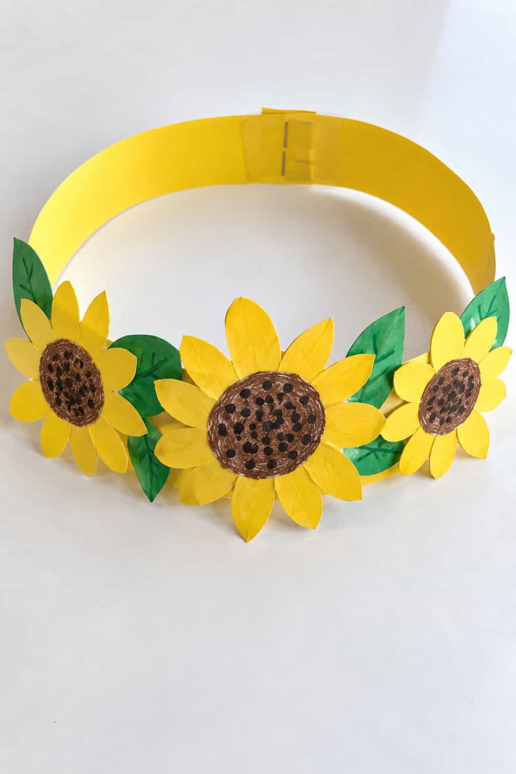 A wearable sunflower crown made from a paper headband curved into a ring, with two or three yellow and brown paper sunflowers standing up along the front and green leaves tucked around them, laid flat on a white table.