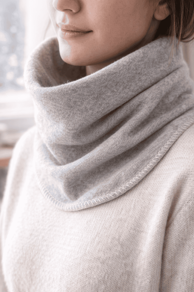 Close-up of a soft oatmeal-colored neck warmer worn over a knit sweater, showing plush fleece texture and delicate hand-stitched detailing along the hem