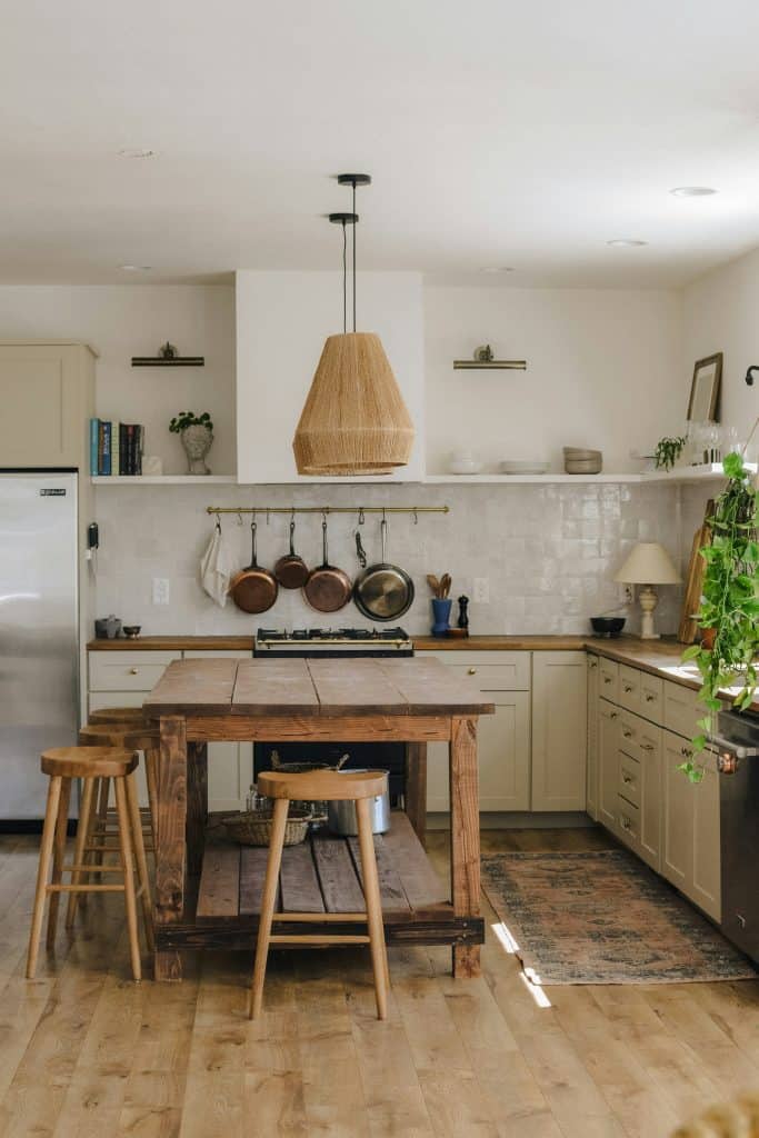 Rustic farmhouse kitchen with wooden island table, woven pendant light, hanging copper pots, open shelving, cream cabinets, and natural wood stools.