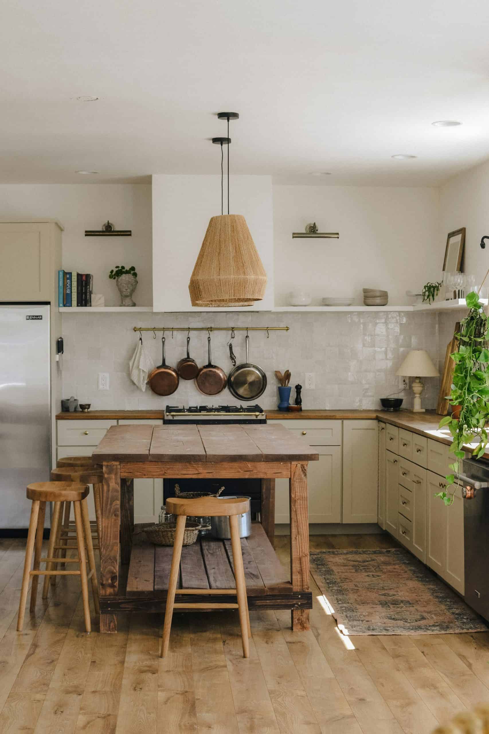 Rustic farmhouse kitchen with wooden island table, woven pendant light, hanging copper pots, open shelving, cream cabinets, and natural wood stools.