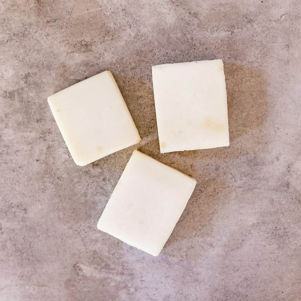 Three square bars of creamy white goat milk soap arranged in a loose triangle on a light stone surface.