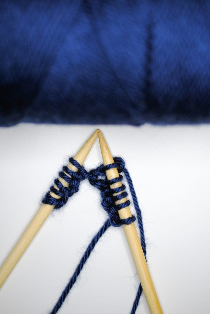 Close-up of two wooden knitting needles crossing as dark blue yarn is worked between them, with several stitches cast on and a strand of yarn trailing below against a white background.