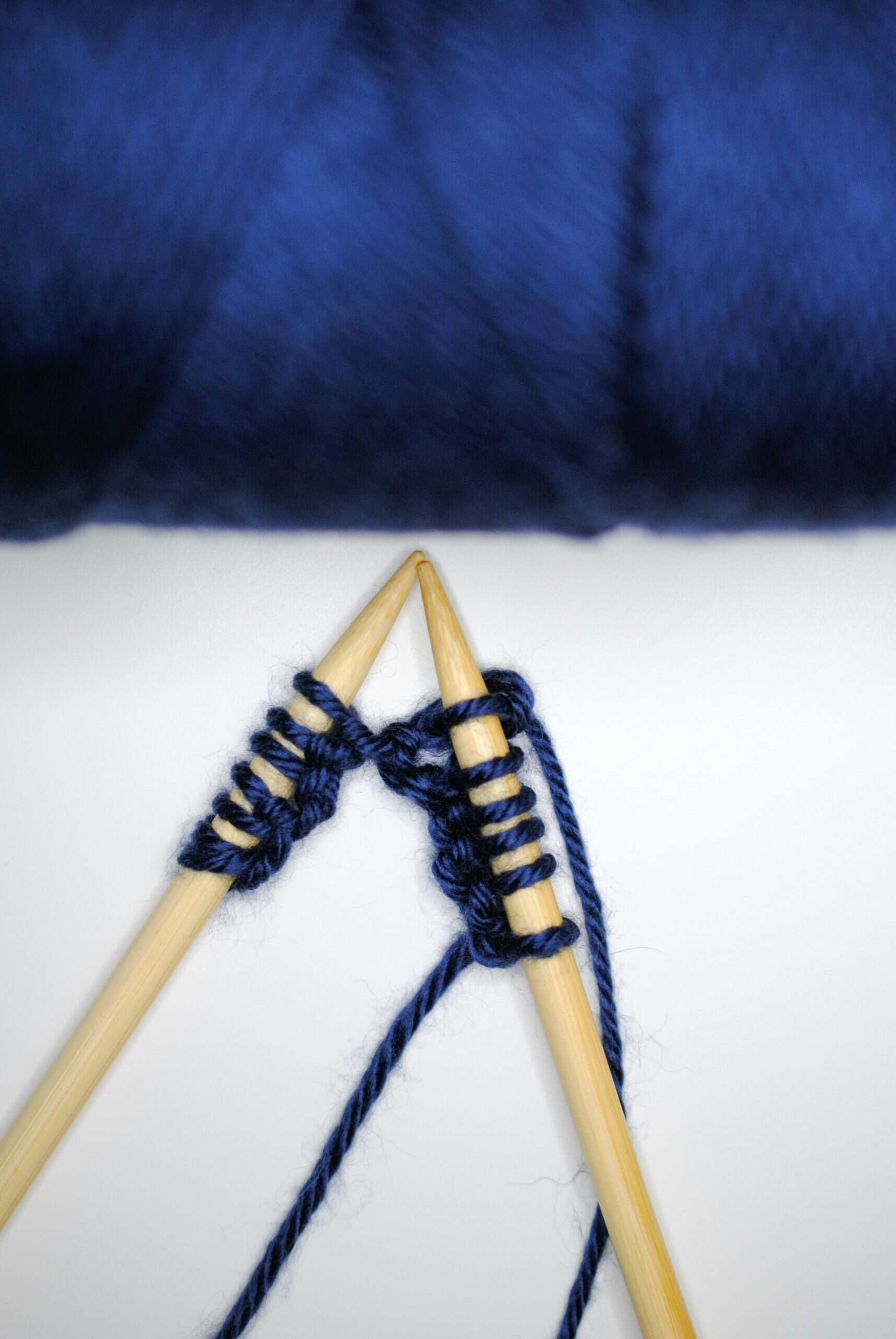 Close-up of two wooden knitting needles crossing as dark blue yarn is worked between them, with several stitches cast on and a strand of yarn trailing below against a white background.