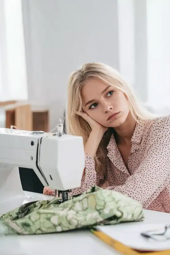 A young woman with long blonde hair wearing a light pink blouse with small black polka dots, sitting at a sewing machine. She rests her head on her hand with a thoughtful, slightly frustrated expression, staring off into the distance.
