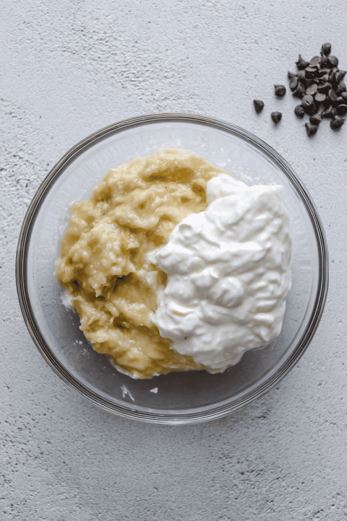 a clear glass mixing bowl containing mashed ripe banana and thick Greek yogurt.