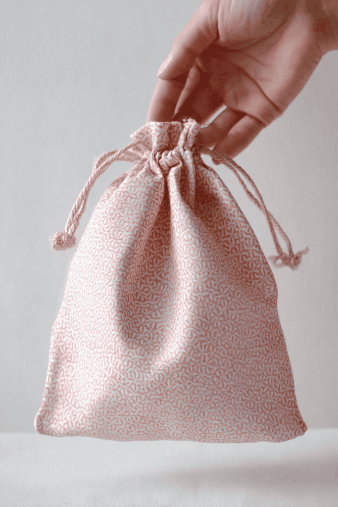 a handmade drawstring fabric bag being gently held up by a human hand against a plain, soft white background