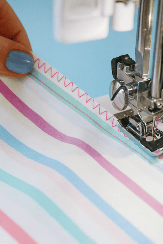 a sewing machine stitching vibrant striped fabric. The sewing machine needle is actively stitching a pink zigzag seam along the fabric edge