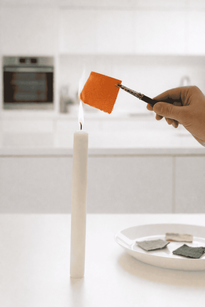 a hand holding a pair of metal tweezers gripping a small fabric swatch directly above the flame of a lit candle during a fabric burn test