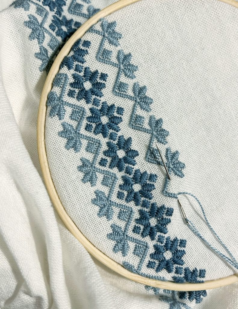 a hand embroidery project in progress, featuring geometric and floral cross-stitch motifs in varying shades of blue