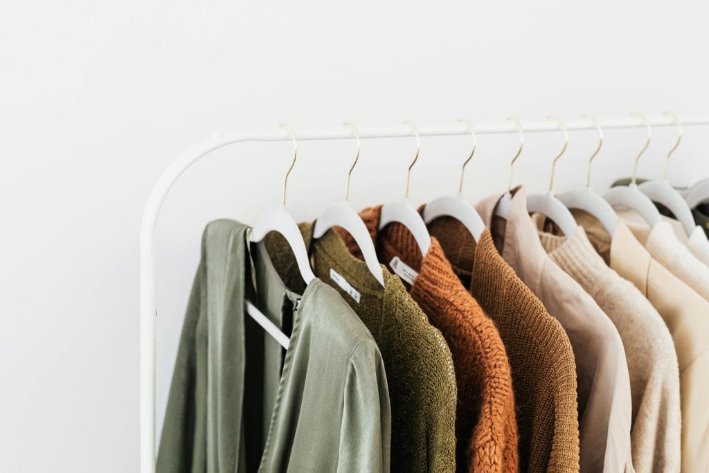 Neutral-toned garments in sage, olive, rust, beige, and cream hanging neatly on white hangers against a minimalist white background.