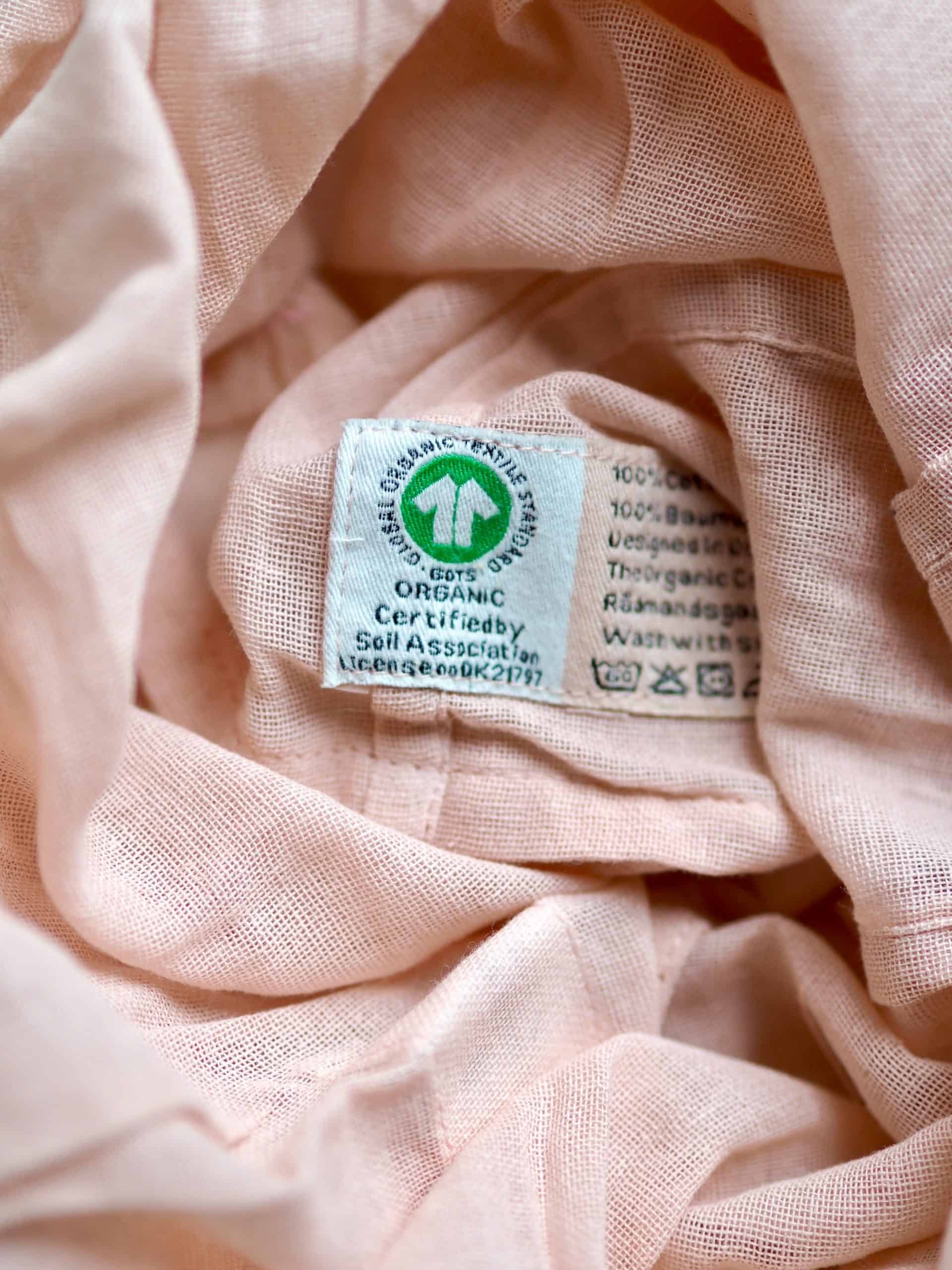 Close-up of a pale blush fabric garment with a visible organic certification label sewn into the seam, surrounded by soft, crumpled folds of lightweight woven fabric.