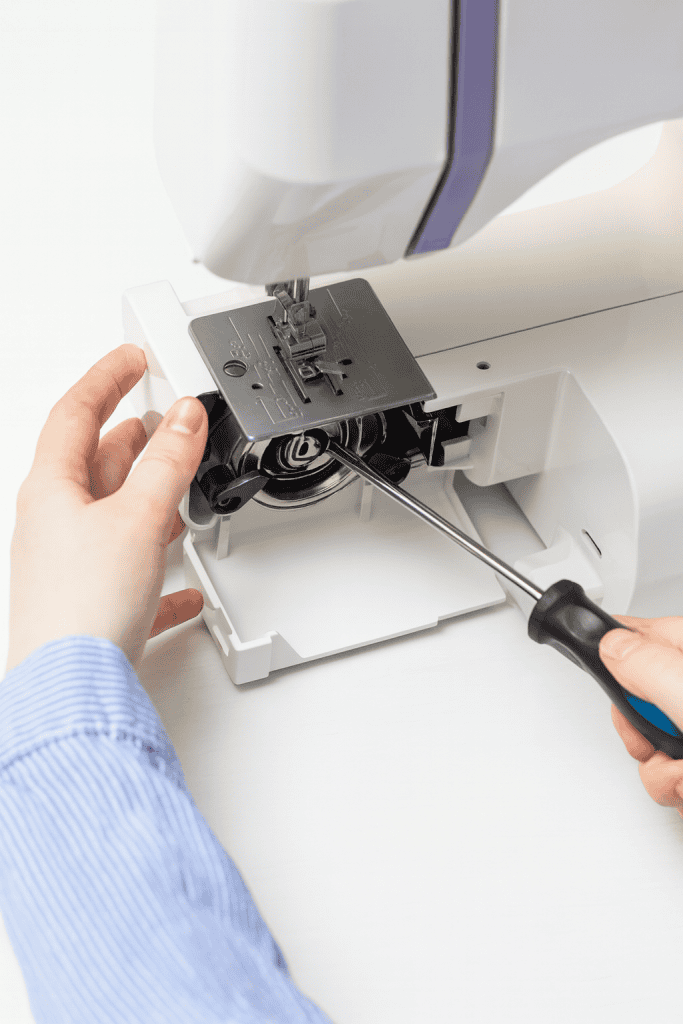 the hands of the individual using a screwdriver to remove or adjust the bobbin case and surrounding components beneath the metal needle plate