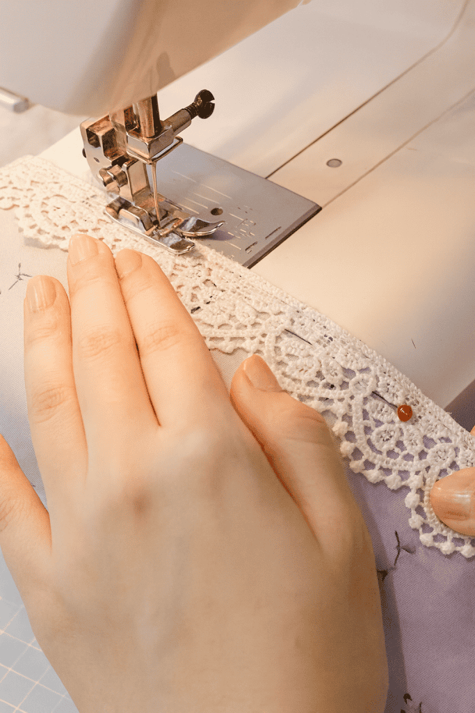 a person sewing white lace trim onto a piece of soft lavender floral fabric using a domestic sewing machine