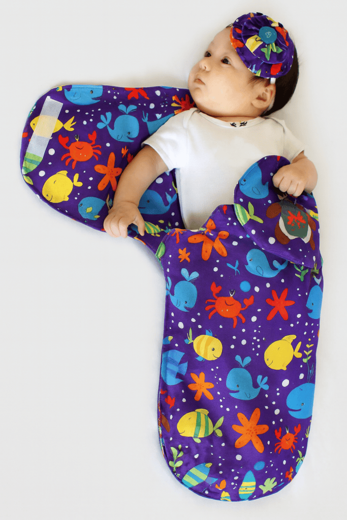 a baby swaddled in a handmade cocoon-style swaddle sack with one arm partially out, displayed on a clean white background.