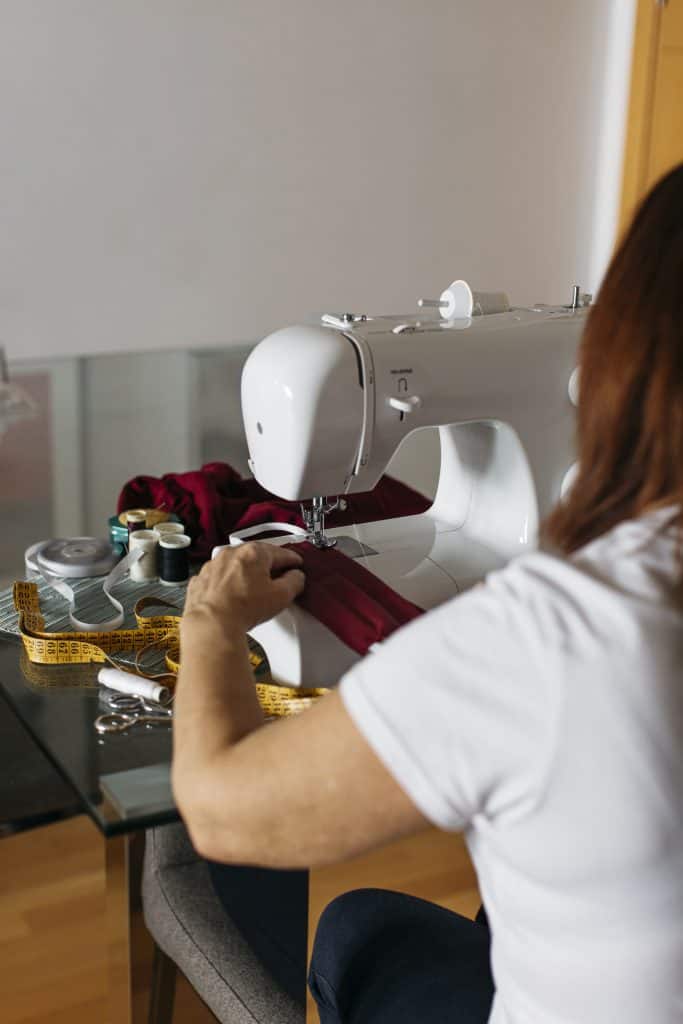 a woman sewing deep red fabric using a modern white sewing machine on a glass-top table in a home setting