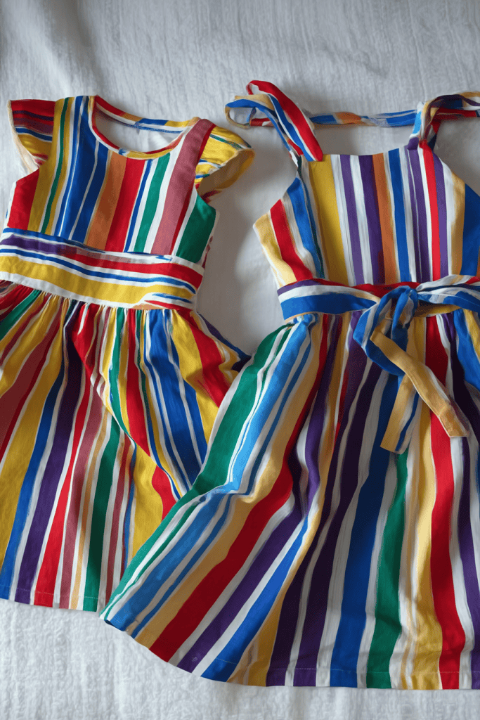 two colorful, handmade girls’ dresses with bold vertical and horizontal multicolor stripe patterns.