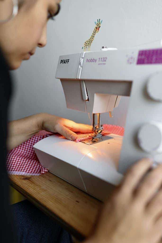 close-up photograph of a person sewing on a white PFAFF Hobby 1132 sewing machine in a cozy indoor workspace