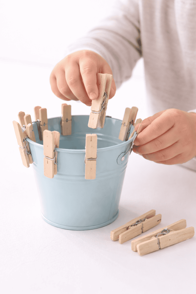 Toddler practicing fine motor skills by clipping wooden clothespins onto a small pastel bucket