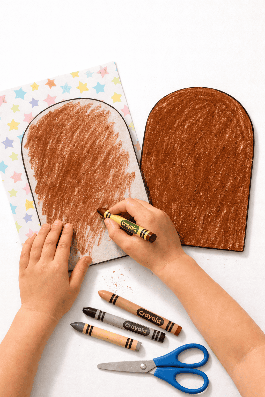 Overhead portrait photo of a child’s hands coloring two rounded-top paper tablets brown with a crayon on a crisp white table.
