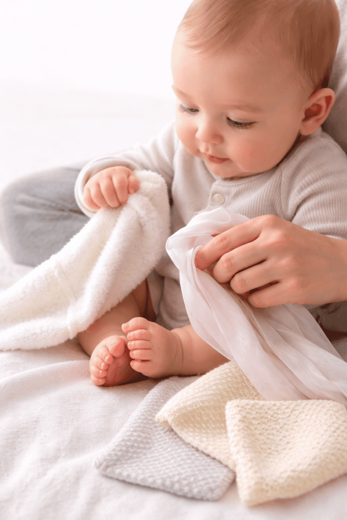 Baby exploring soft fabrics while seated on a caregiver’s lap, gently grasping a plush white cloth and silky scarf