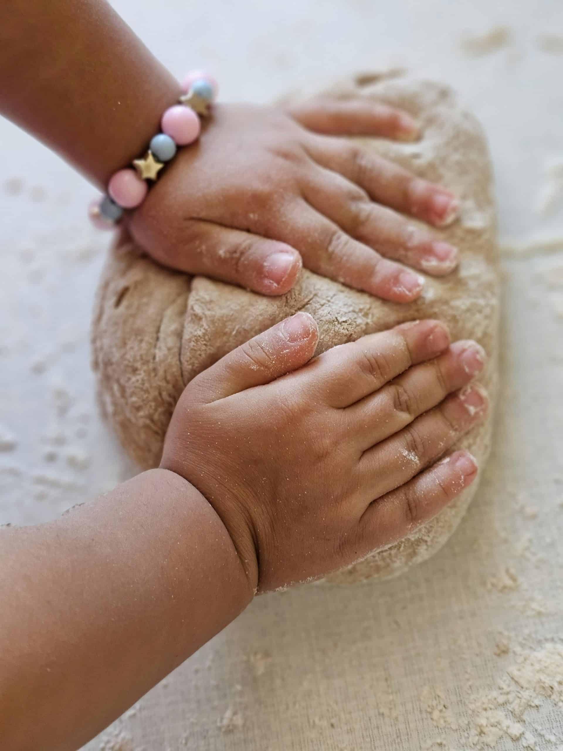 a small child’s hands kneading a rustic ball of bread dough on a lightly floured surface