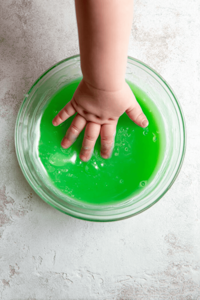 a child’s hand submerged in a bowl of bright green oobleck (a non-Newtonian fluid made from cornstarch and water).