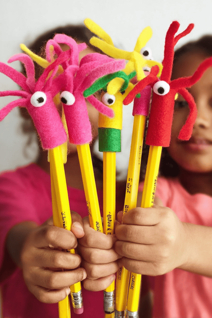 a group of children's hands holding bright yellow pencils, each topped with a handmade felt pencil topper