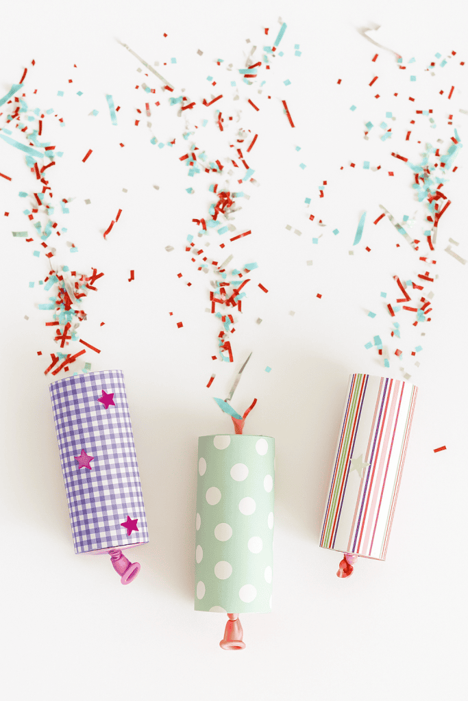 three festive handmade confetti poppers mid-burst, ejecting colorful metallic confetti into the air