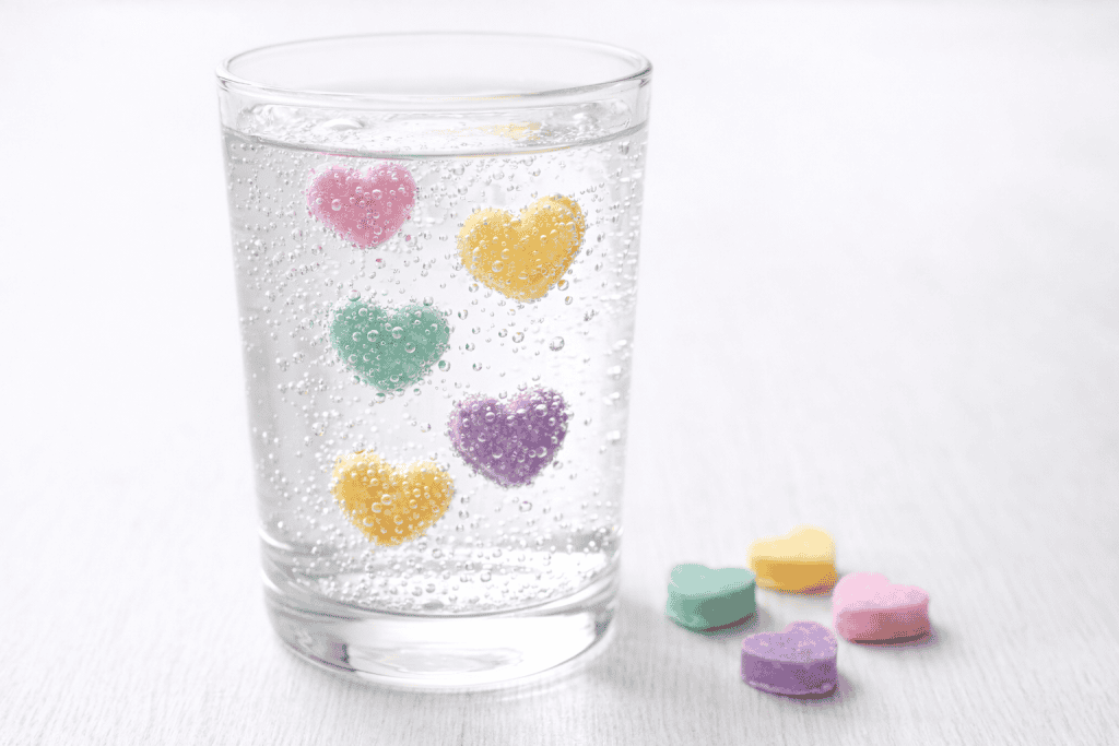 a clear glass filled with fizzy clear soda (like Sprite) on a simple white surface. Inside the glass, several pastel conversation candy hearts (pink, yellow, green, purple) are visibly suspended at different heights, with tiny carbonation bubbles clinging to their surfaces.