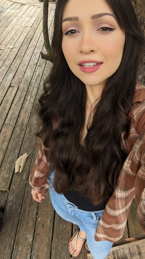 Woman taking a top-down selfie on a wooden deck, wearing a brown plaid shirt, black top, jeans, and sandals.
