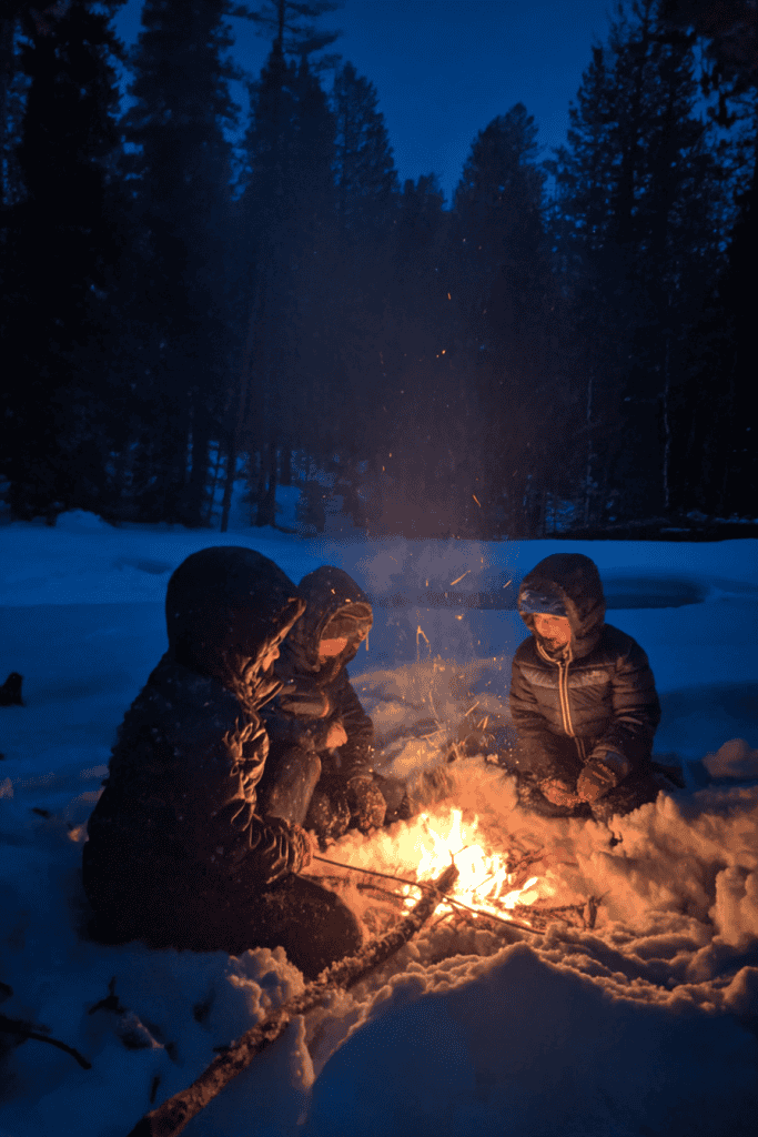 children gathered around a small campfire in the snow under a darkening twilight sky