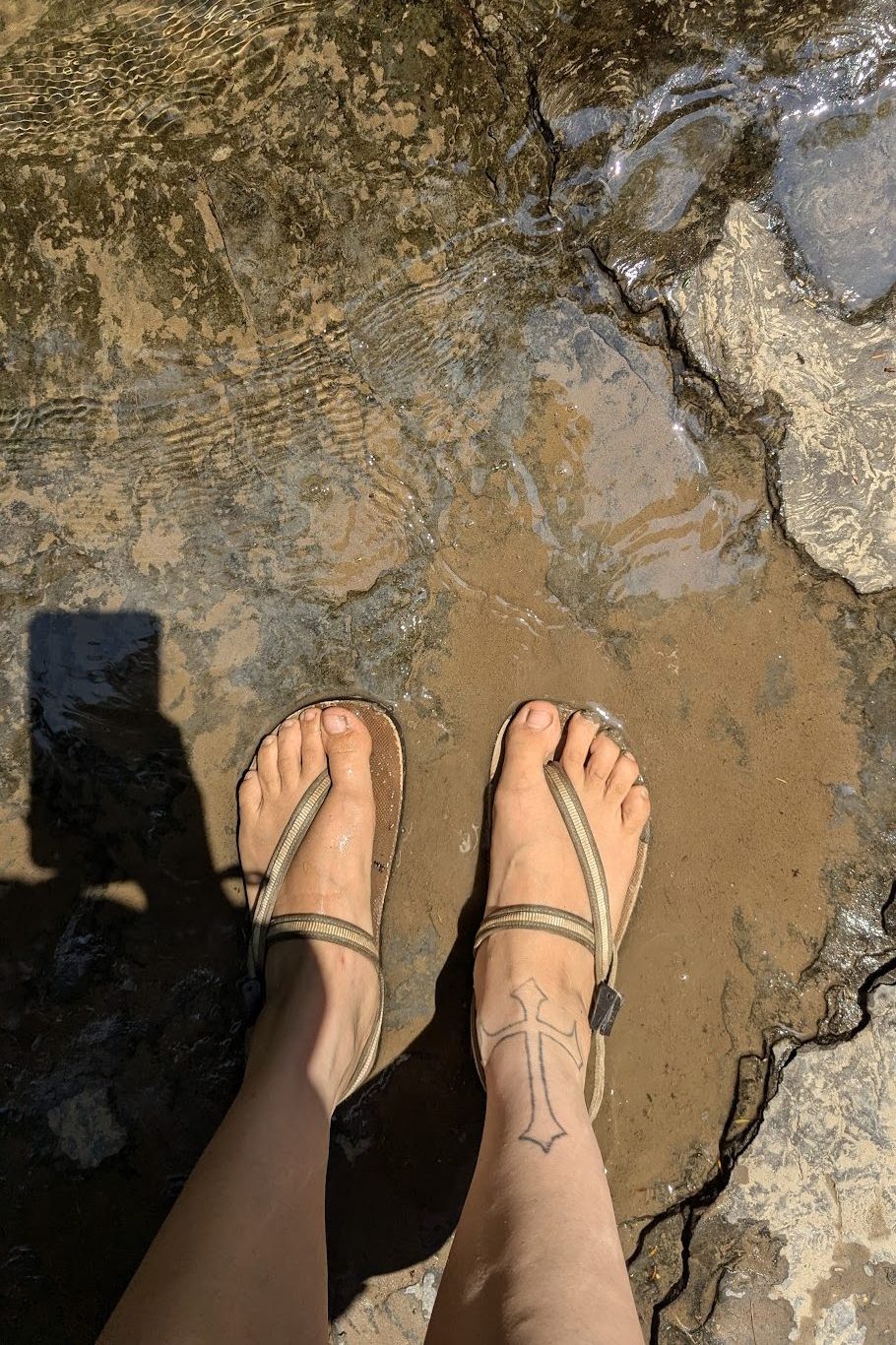 a person standing barefoot in thin-strapped sandals on wet, sunlit rocks partially submerged in shallow, clear stream water.