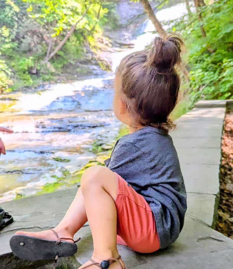 a young child sitting on the edge of a stone ledge in a serene forest setting, overlooking a shallow cascading stream.