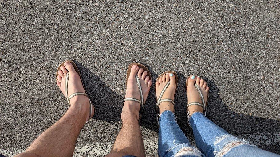 a couple standing side-by-side on a sunlit asphalt surface, both wearing minimalist, earth runner sandals.