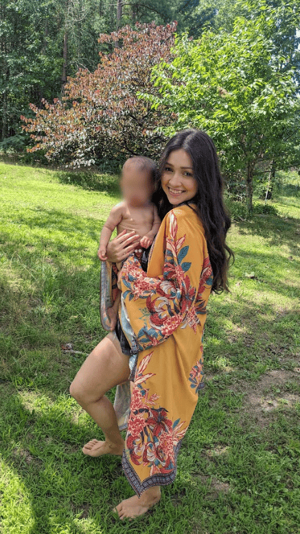 Woman smiling and holding a baby outdoors on green grass, wearing a yellow floral kimono, barefoot in a sunny garden setting.