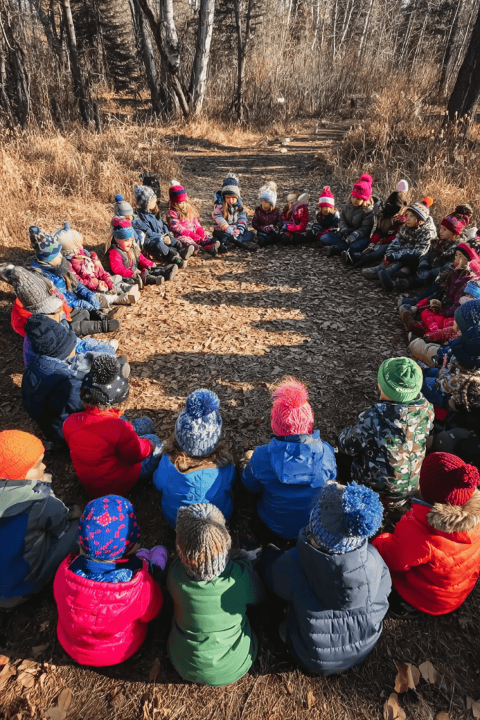 a large group of young children and two adult educators sitting in a perfect circle outdoors on a forest path during late autumn.