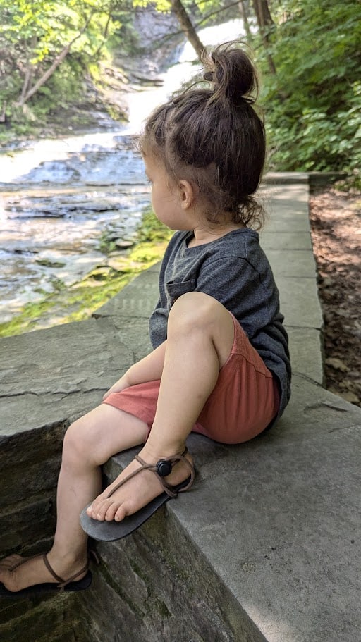 a young child sitting on the edge of a stone ledge in a serene forest setting, overlooking a shallow cascading stream.