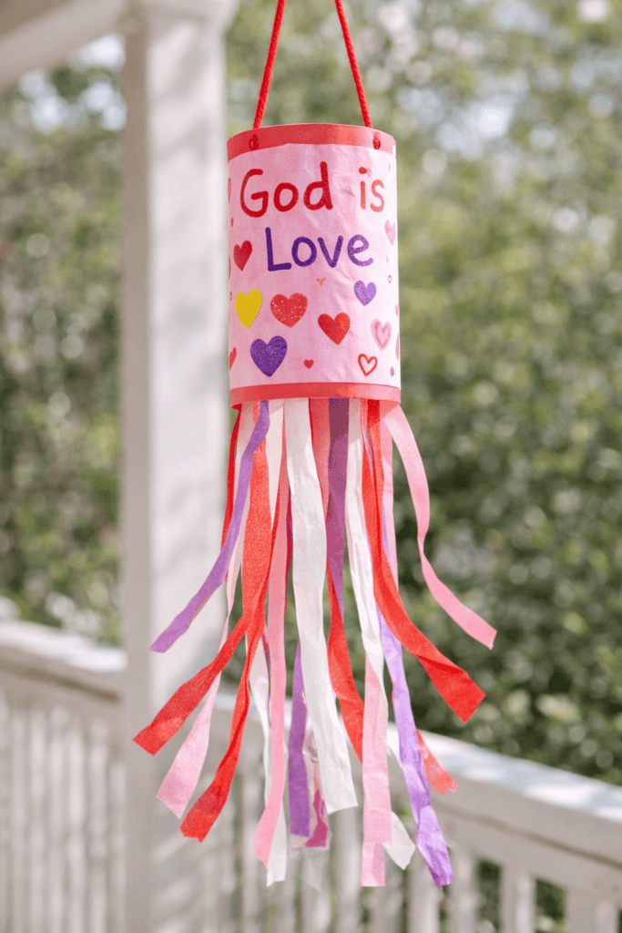a handmade “God Is Love” Valentine windsock hanging gently outdoors on a porch or from a tree branch.