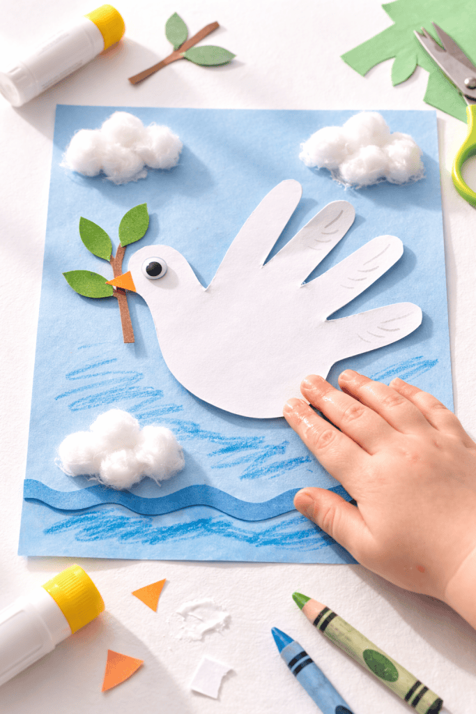 Toddler-made white handprint dove holding an olive branch on blue paper with cotton ball clouds
