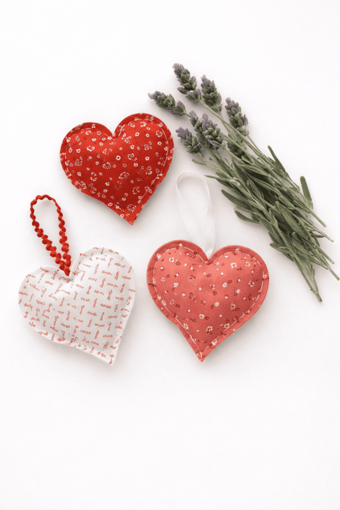three handmade heart-shaped lavender sachets, crafted from soft cotton fabric in warm shades of red, coral, and cream