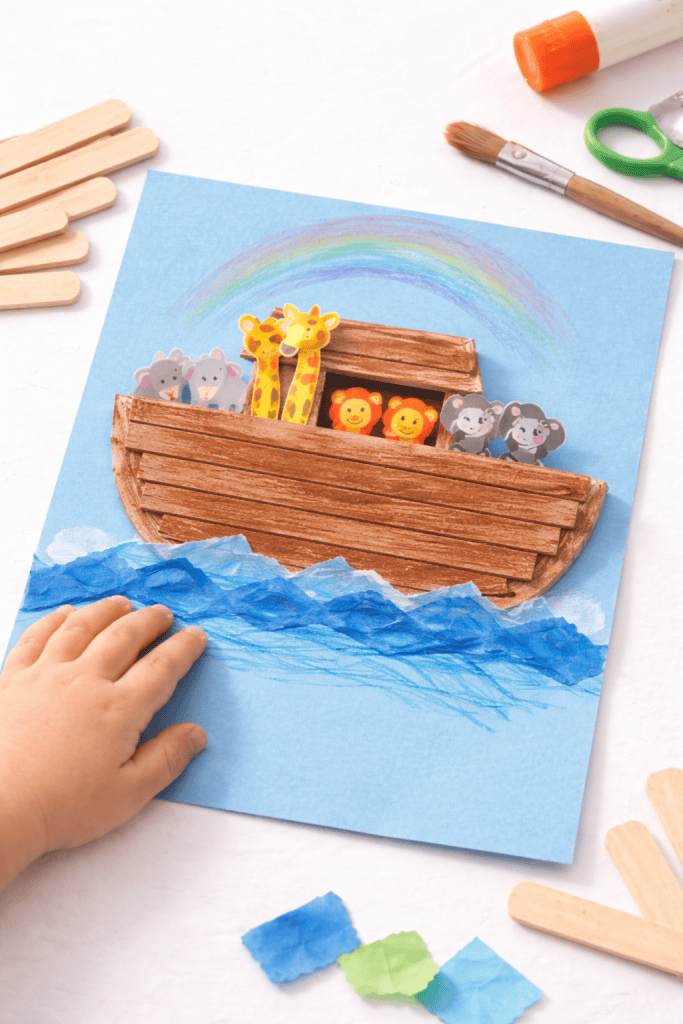 Toddler-made popsicle stick Noah’s Ark craft on blue construction paper, decorated with animal stickers and tissue paper waves